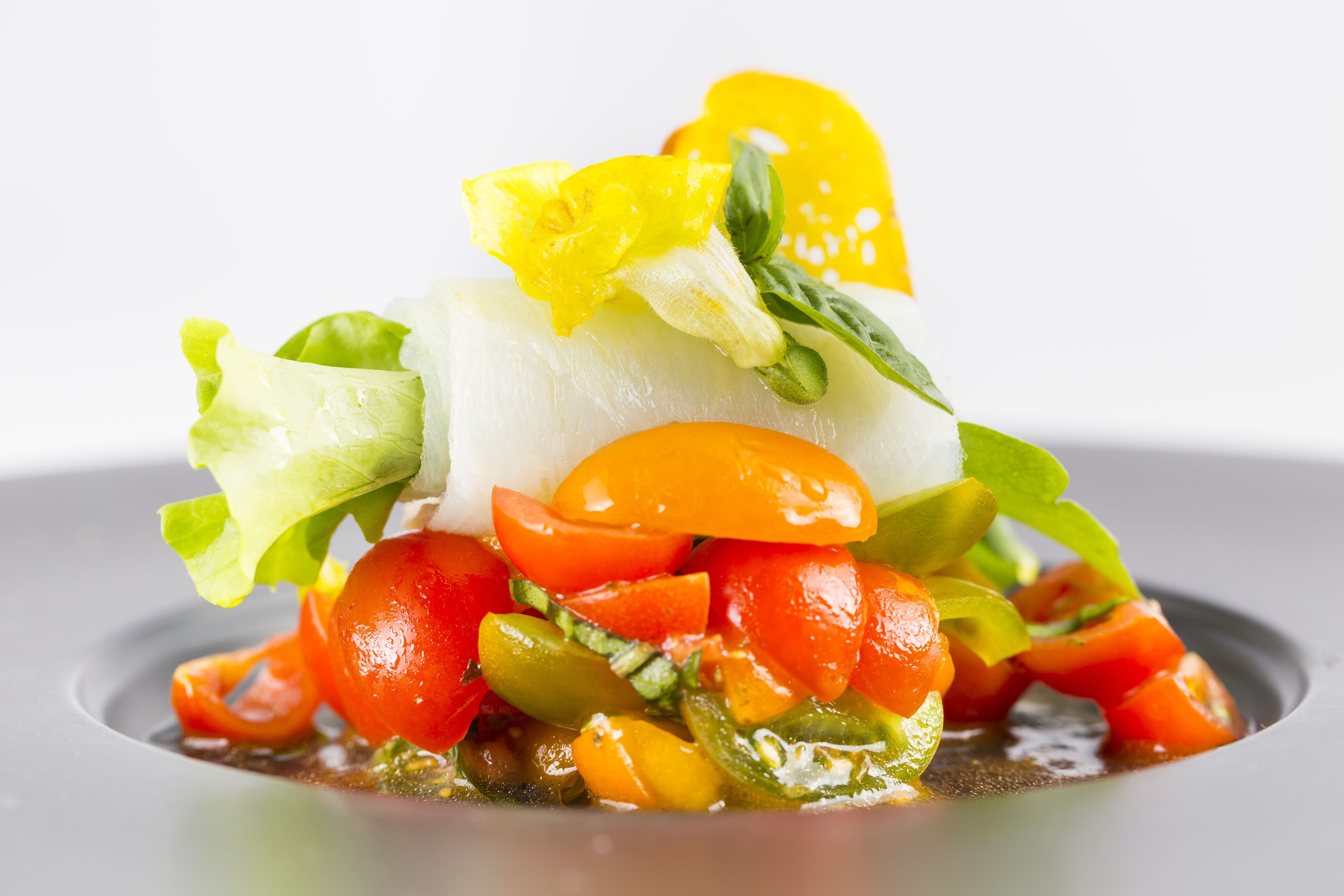 Un plato con un pescado blanco como pieza central, rodeado de verduras coloridas y adornado con hojas verdes y pétalos de flores amarillas.