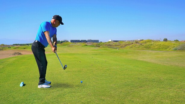 Un terrain de golf sous un ciel dégagé, une personne vêtue d’une chemise bleue et d’un pantalon noir se préparant à frapper une balle de golf, et un bâtiment en arrière-plan.