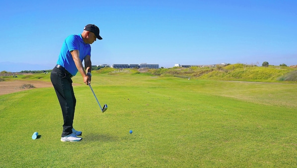 A golf course with a clear sky, a person in a blue shirt and black pants preparing to hit a golf ball, and a building in the background.