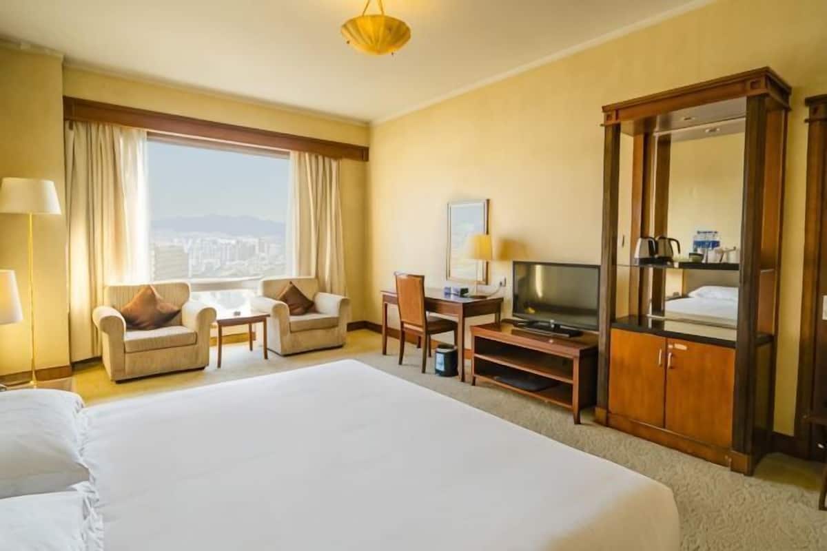 Une chambre d’hôtel avec un grand lit, un bureau avec une télévision, une chaise, un canapé, une armoire et une grande fenêtre avec des rideaux.