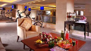 Un vestíbulo de hotel con un sofá, una mesa de centro con frutas y revistas, y bicicletas estáticas al fondo.