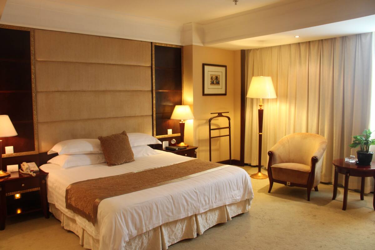Quarto de hotel com uma cama grande, duas mesas de cabeceira com luminárias, uma cadeira, uma mesinha com uma planta e um quadro na parede.