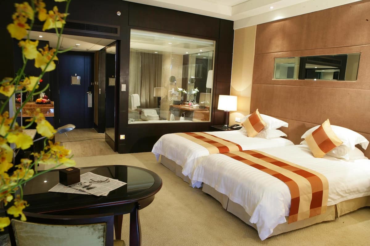 Habitación de hotel con una cama grande, un escritorio con silla, un espejo y un baño visible a través de una puerta de cristal.