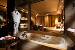 Een luxe hotelkamer met een groot bad vol rozenblaadjes, een badjas hangend aan een deur en een bed met uitzicht op de kamer.