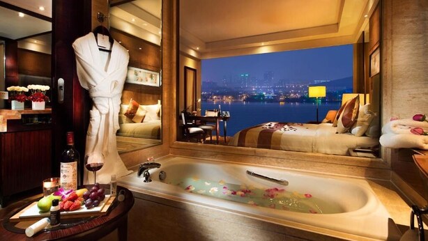 A luxurious hotel room with a large bathtub filled with rose petals, a city view, and a robe hanging on the door. - New Century Grand Hotel Xuzhou (Xuzhou)