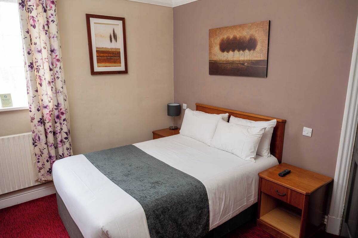 A neatly made bed with a grey blanket, a bedside lamp, and a remote control on a wooden nightstand.