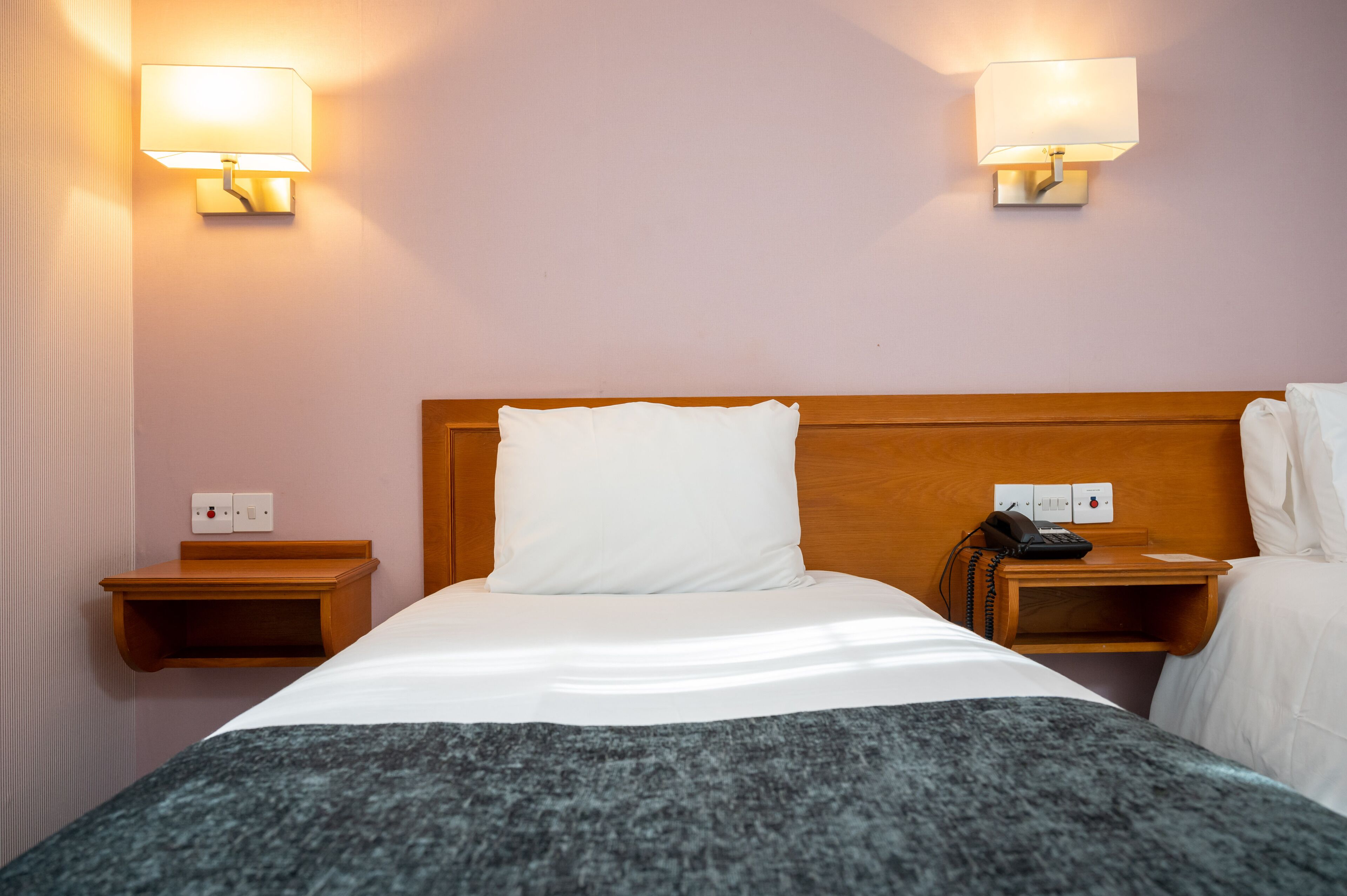 A hotel room with a wooden headboard, a white bedspread, a gray throw blanket, two bedside tables with lamps, and a telephone.