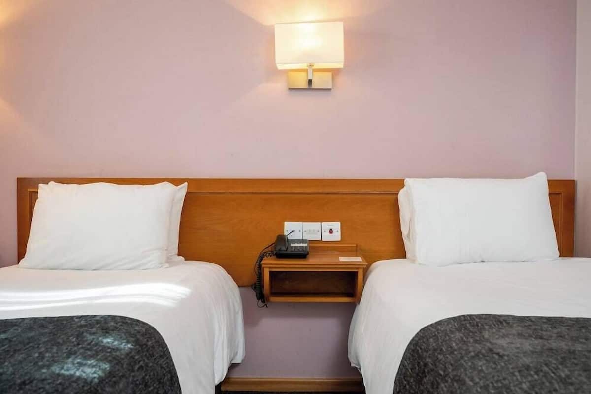 A hotel room with two beds, a bedside table with a lamp, and a wall-mounted light fixture.