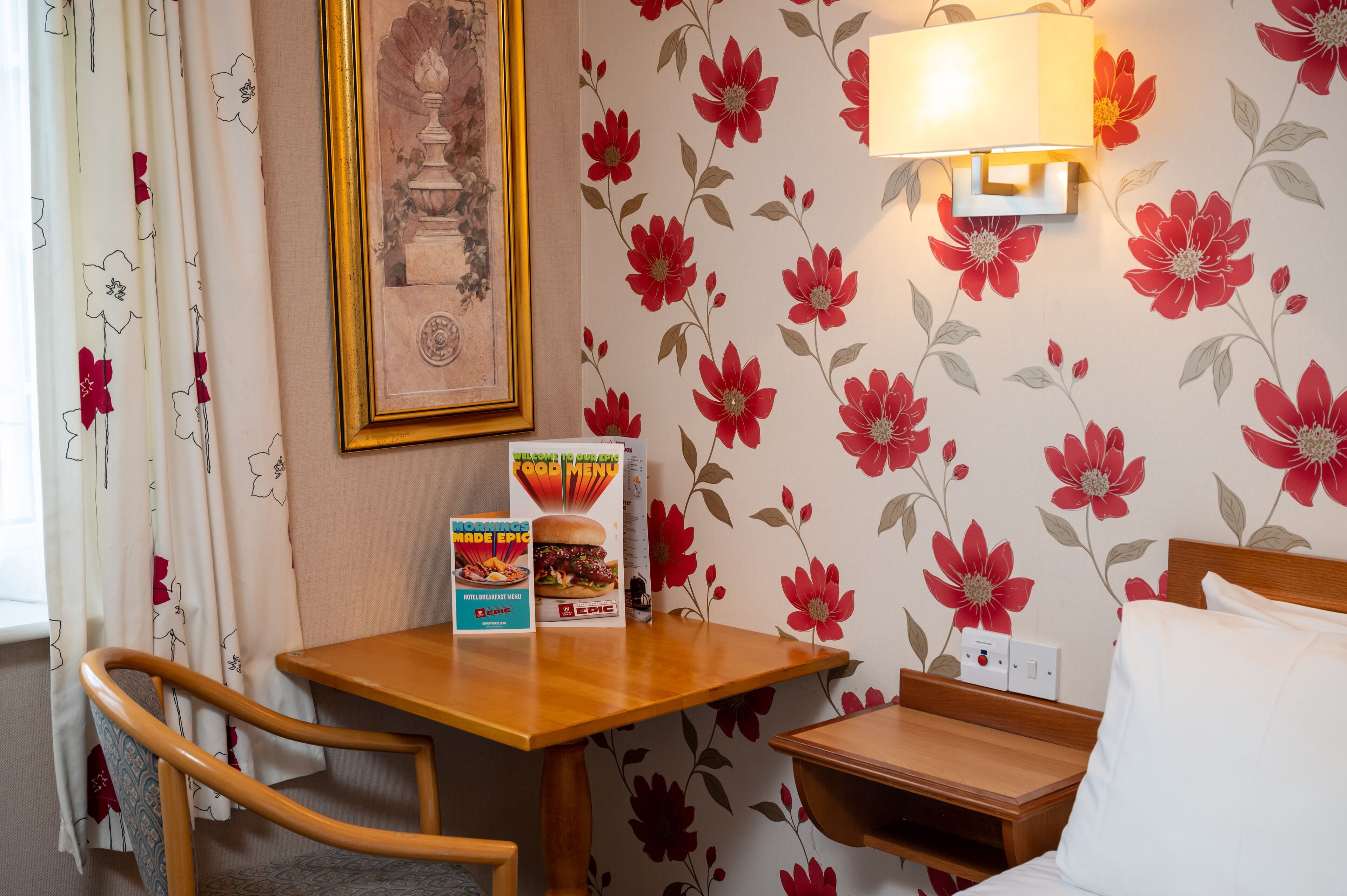 A hotel room with a wooden table, a framed picture on the wall, a floral wallpaper, and a breakfast menu on the table.
