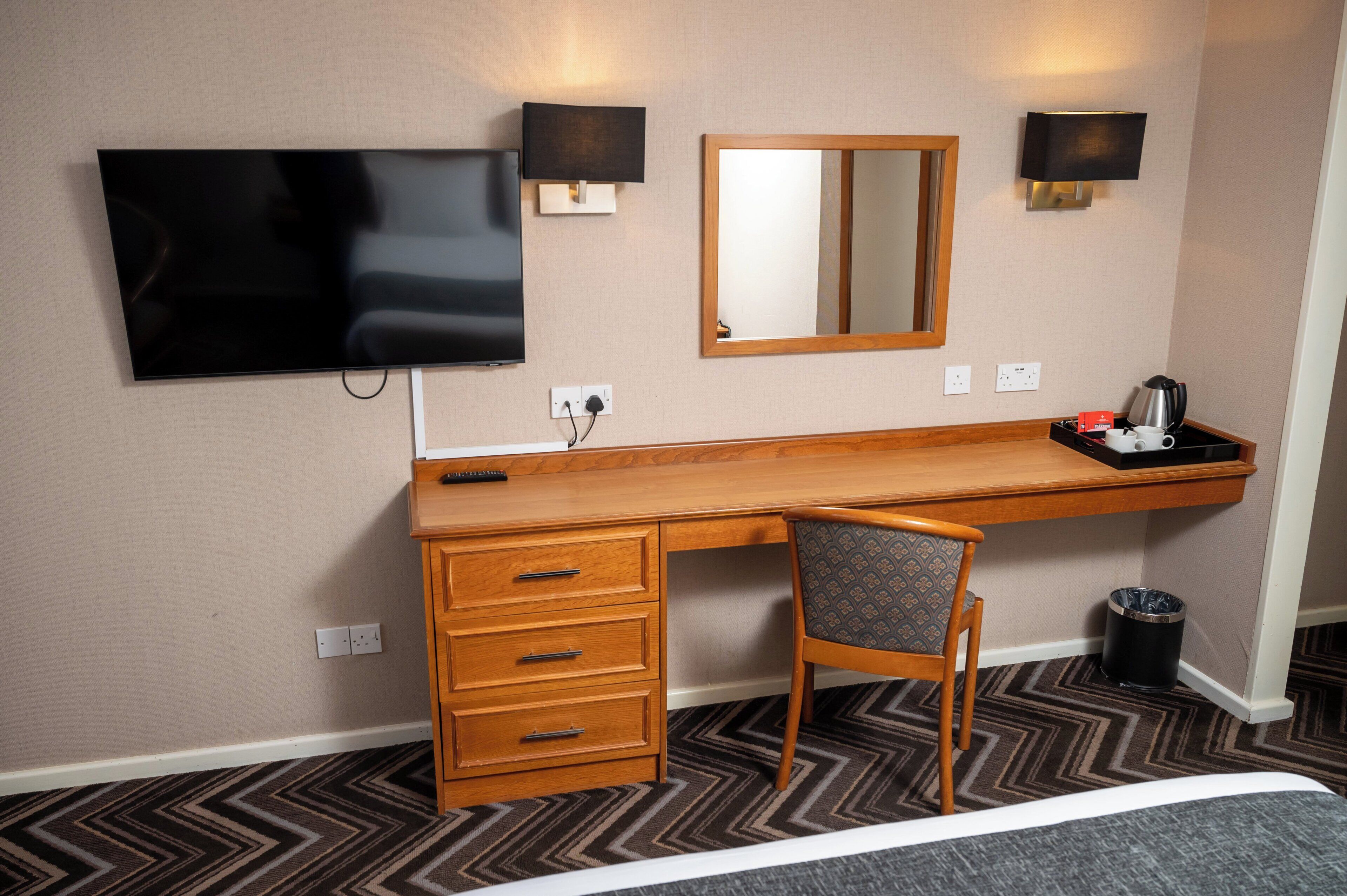 A hotel room with a wooden desk, a chair, a television mounted on the wall, a mirror, and a small table with a coffee pot.