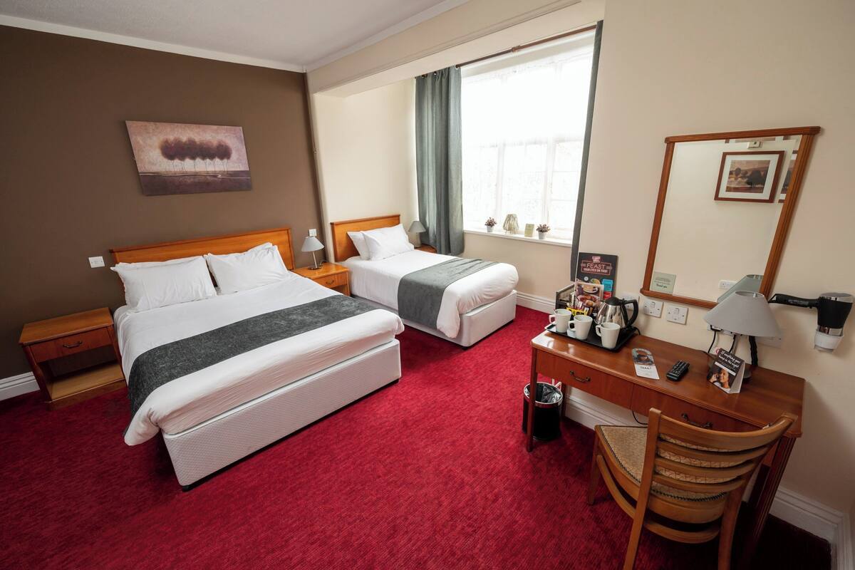 A hotel room with two beds, a desk with a lamp, a chair, a small trash bin, and a window with curtains.
