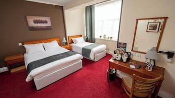 Een hotelkamer met twee bedden, een bureau met lamp, een stoel, een kleine prullenbak en een raam met gordijnen.