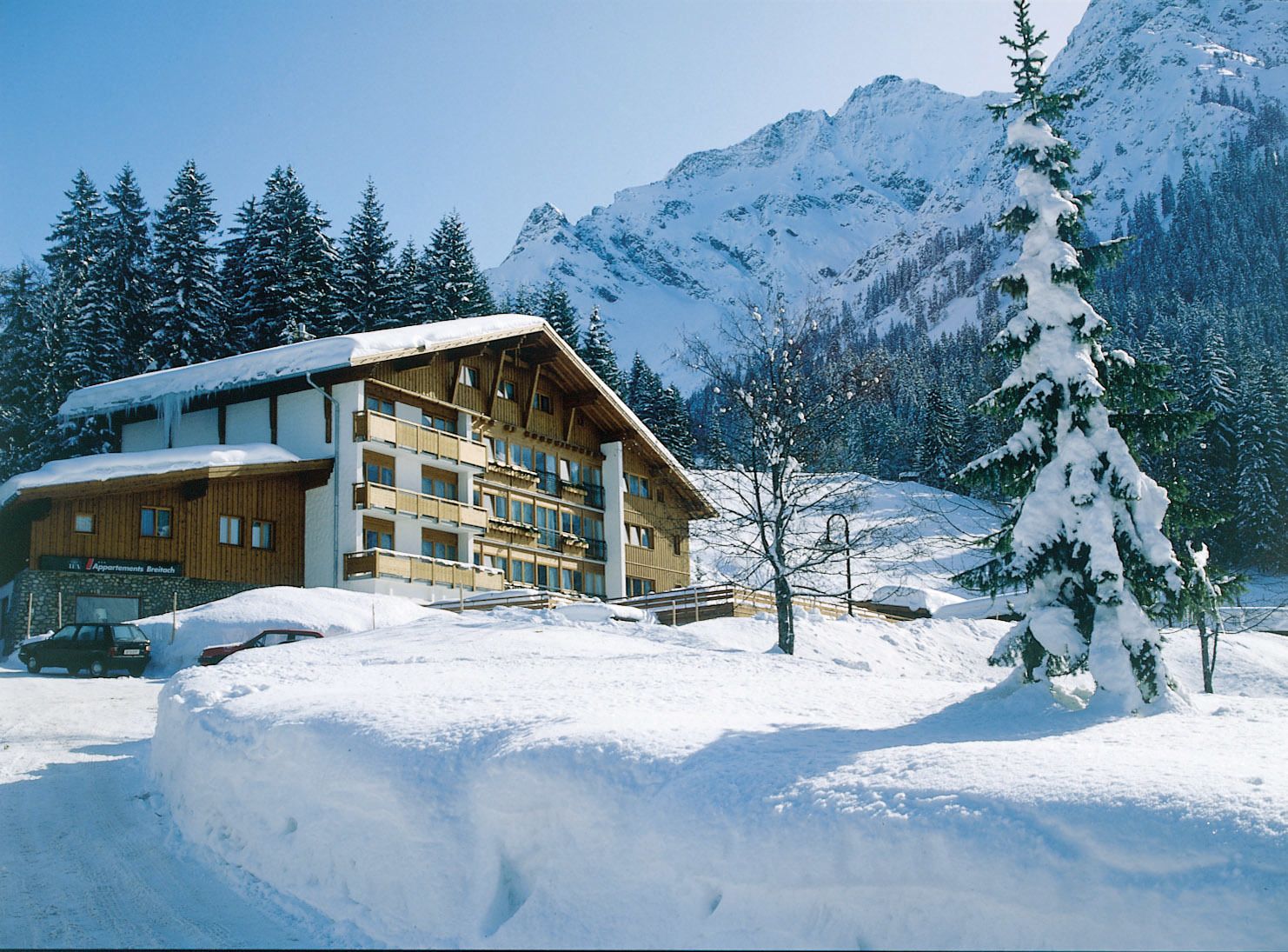 A wooden chalet with multiple balconies, surrounded by snow-covered mountains and trees.