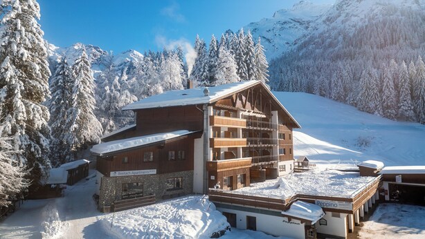 A wooden building with multiple balconies, surrounded by snow-covered trees and mountains.