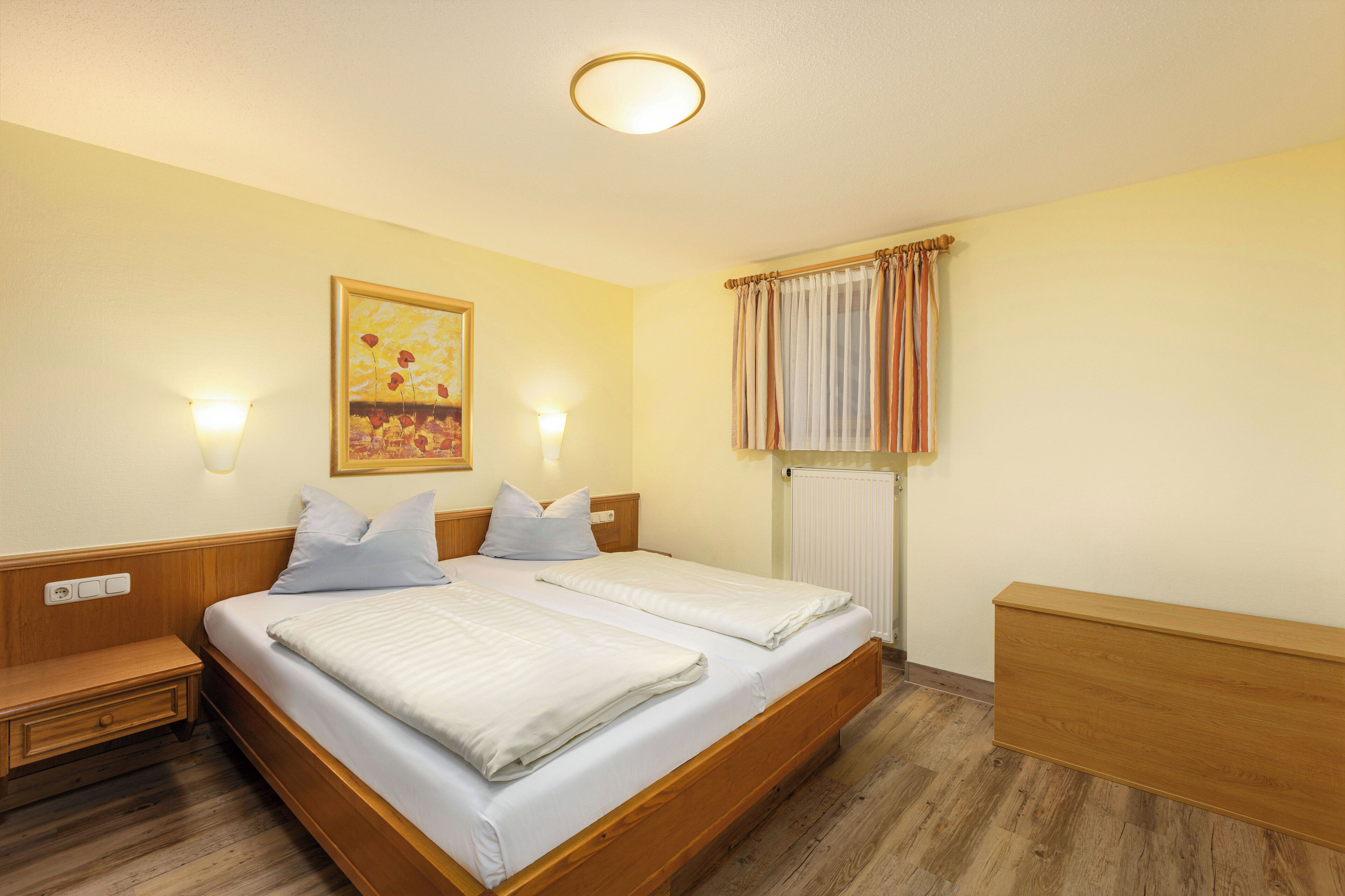 A double bed with white linens, two bedside tables, a wall-mounted light, a painting, and a window with curtains.