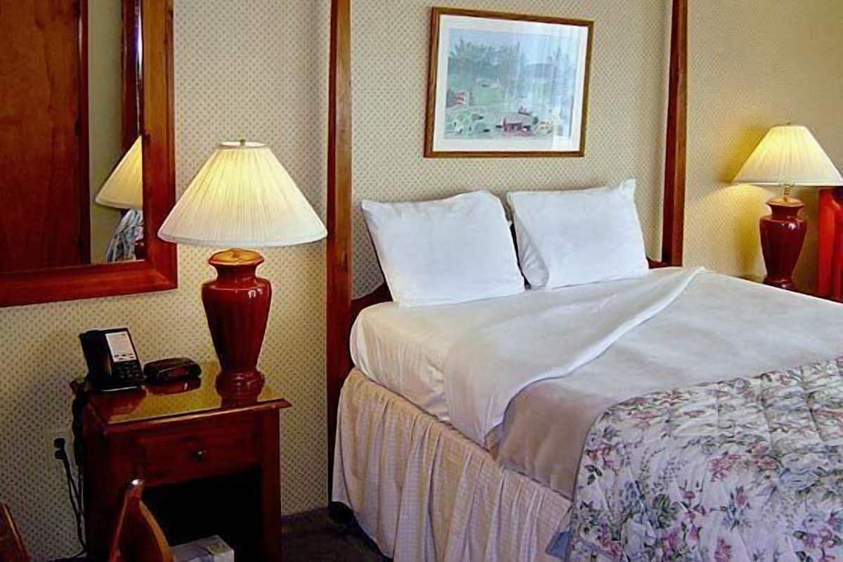 A hotel room with a four-poster bed, two bedside lamps, a nightstand with a telephone and a red vase, and a framed picture on the wall.