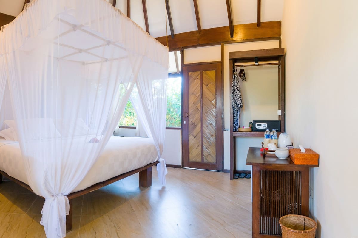 Un dormitorio con cama con dosel, una mesita de noche con lámpara, suelo de madera, un baño con espejo y una pequeña mesa con artículos de aseo.