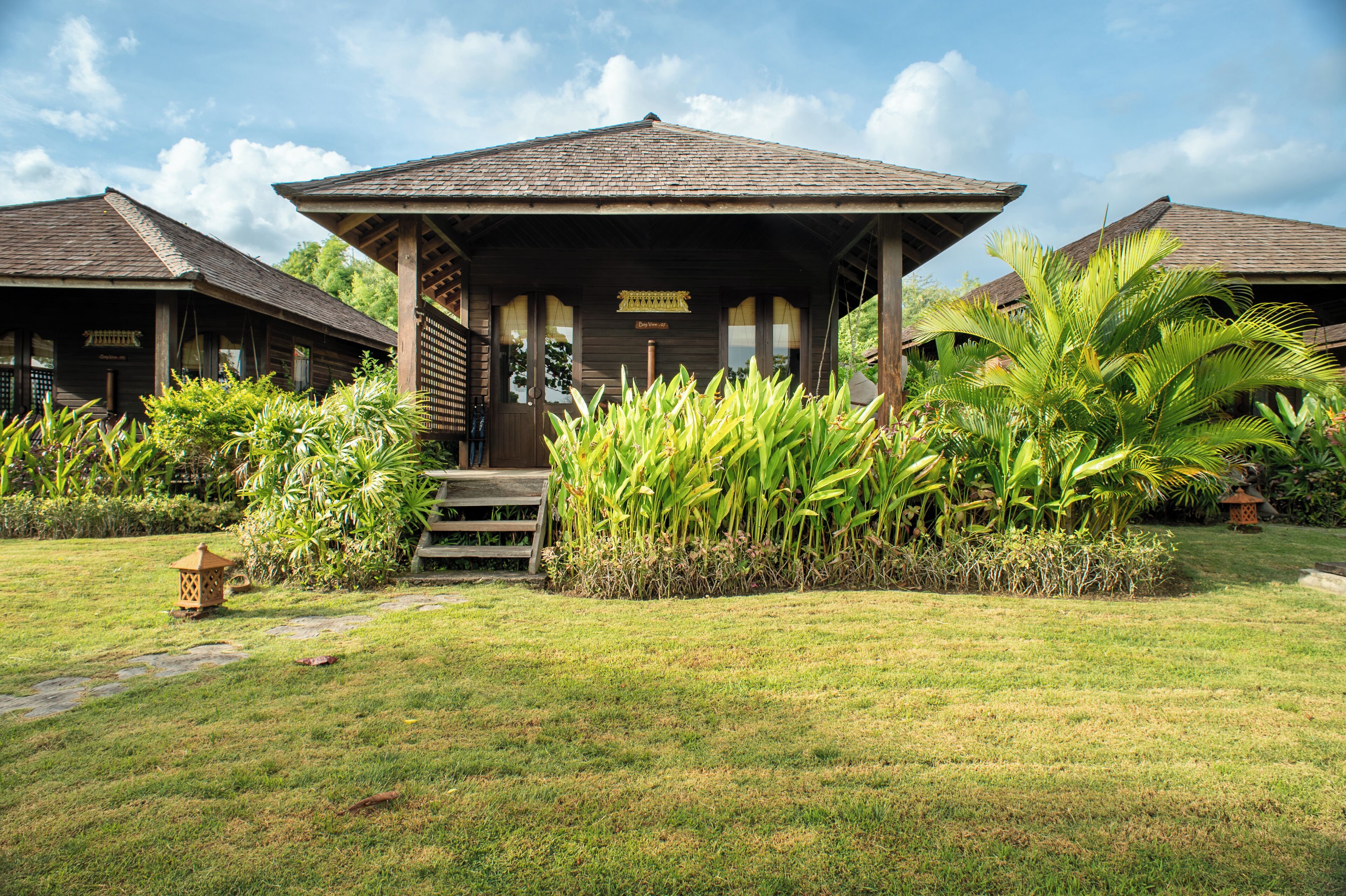 Un bâtiment en bois doté d’un porche, entouré de plantes tropicales et surplombé d’un ciel dégagé.
