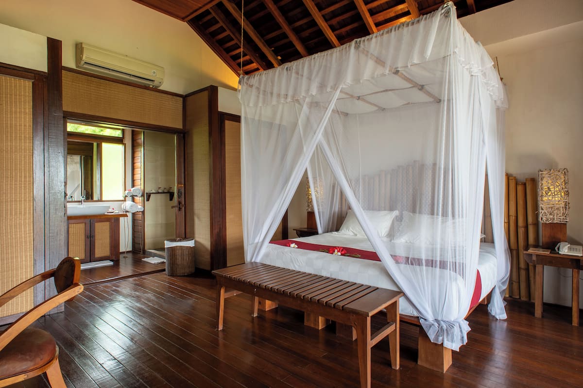 Una habitación con piso de madera, una cama con dosel, un banco de madera, una silla, una mesita de noche y un espejo.