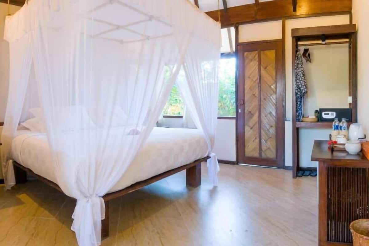 Habitación con cama bajo dosel, banca de madera, una mesita con artículos de tocador y puerta que da al exterior.
