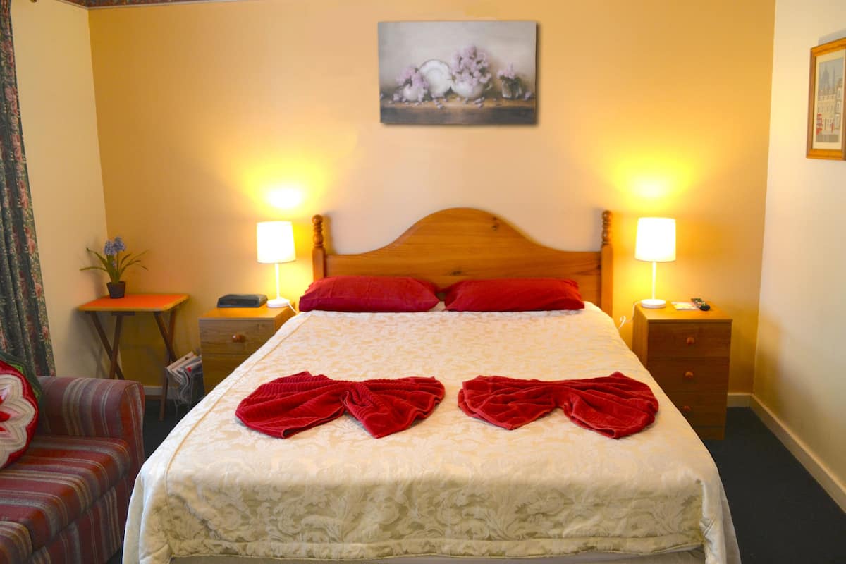 A neatly made bed with red towels, two bedside lamps, a wooden headboard, a small table with a plant, and a framed picture on the wall.