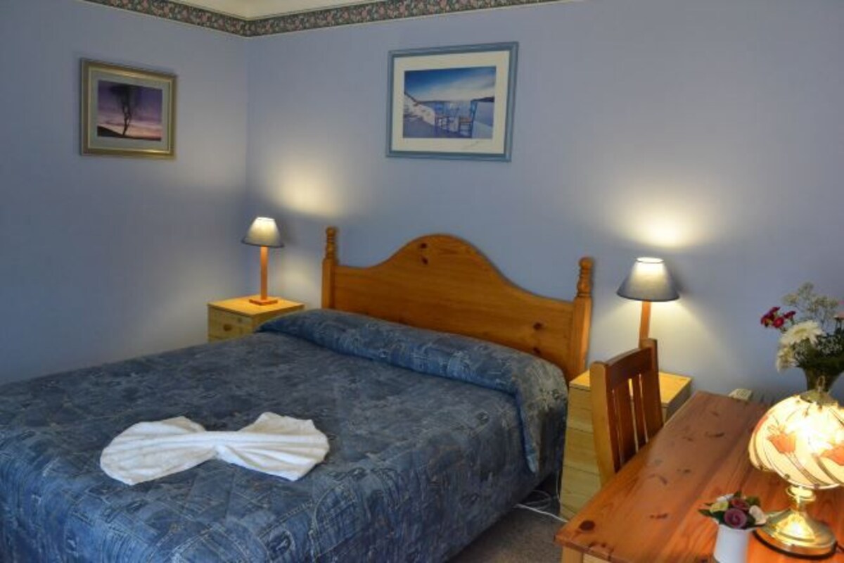 A bedroom with a bed, two bedside tables with lamps, a wooden headboard, and two framed pictures on the wall.