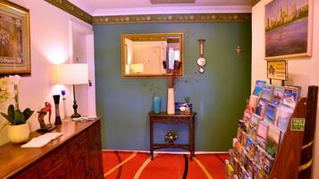 A hotel lobby with a red carpet, a wooden reception desk, a wall-mounted clock, a framed mirror, and a display of travel brochures.