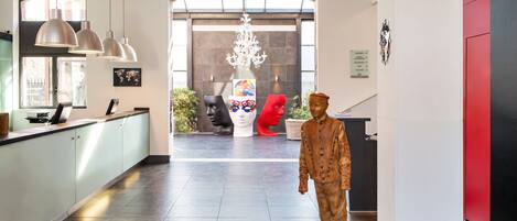 A modern hotel lobby with a reception desk, pendant lights, a large chandelier, and a statue of a person in traditional attire.