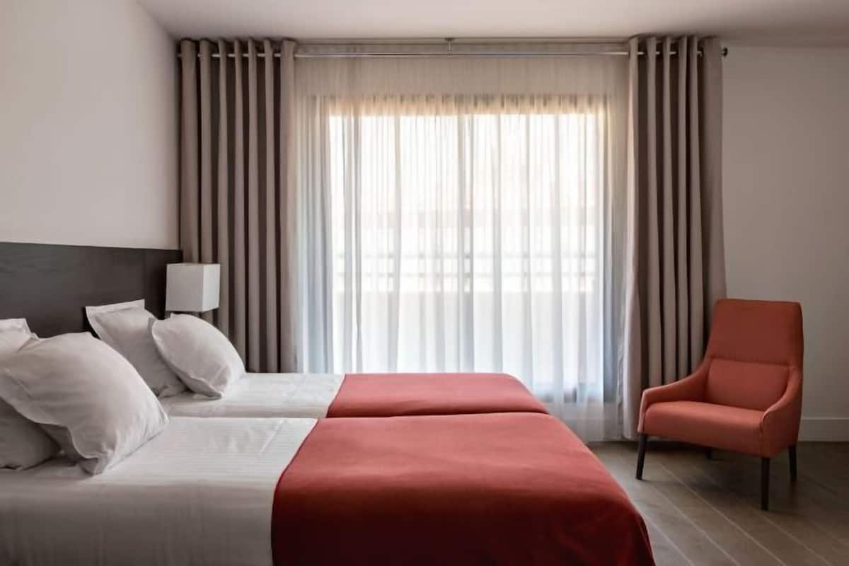 Hotellihuone, jossa on kaksi sänkyä, punainen penkki, tuoli ja ikkuna, jossa on verhot.