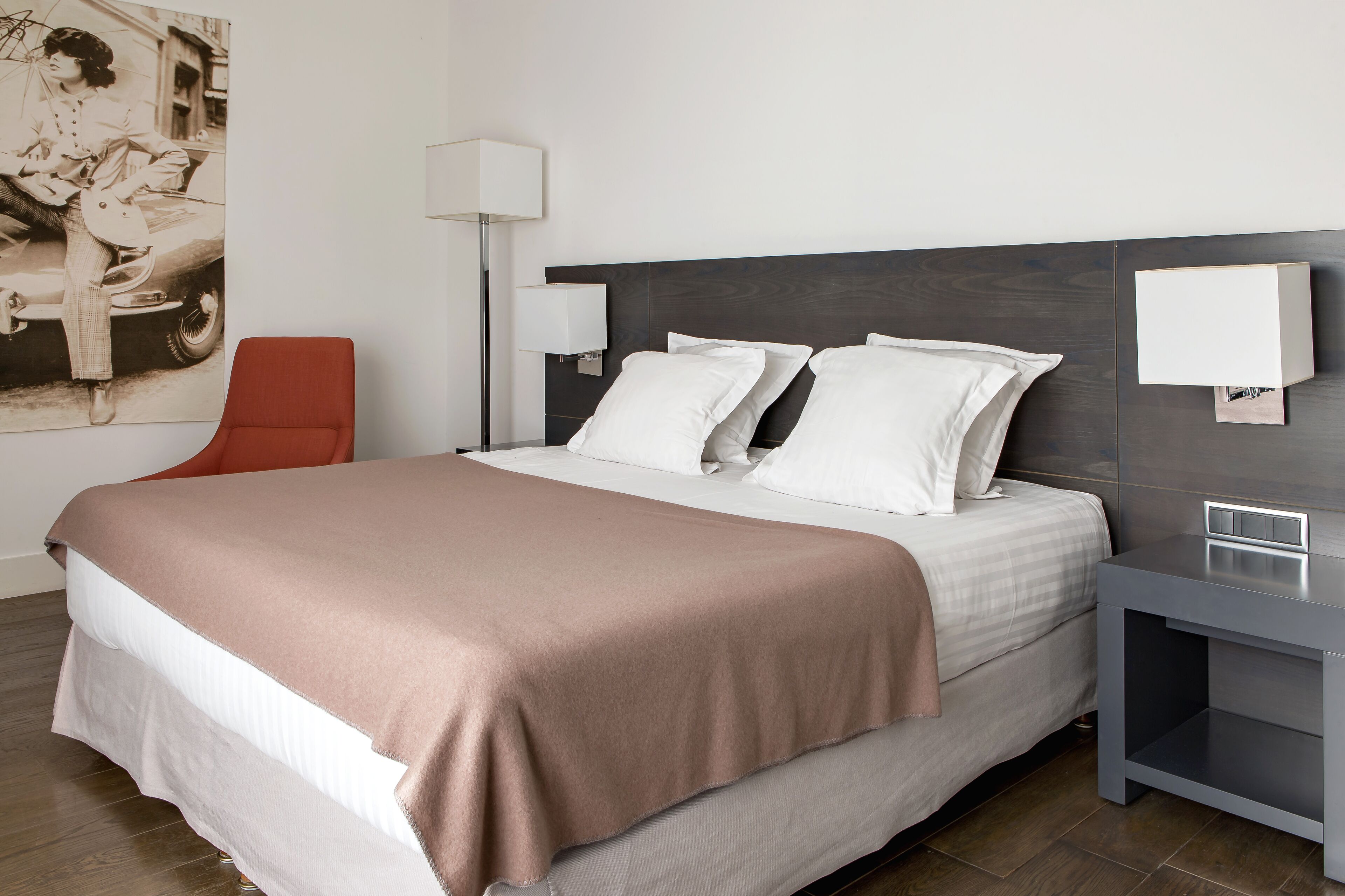 Una habitación de hotel moderna con una cama grande, dos mesitas de noche, una silla roja y una lámpara de pared.