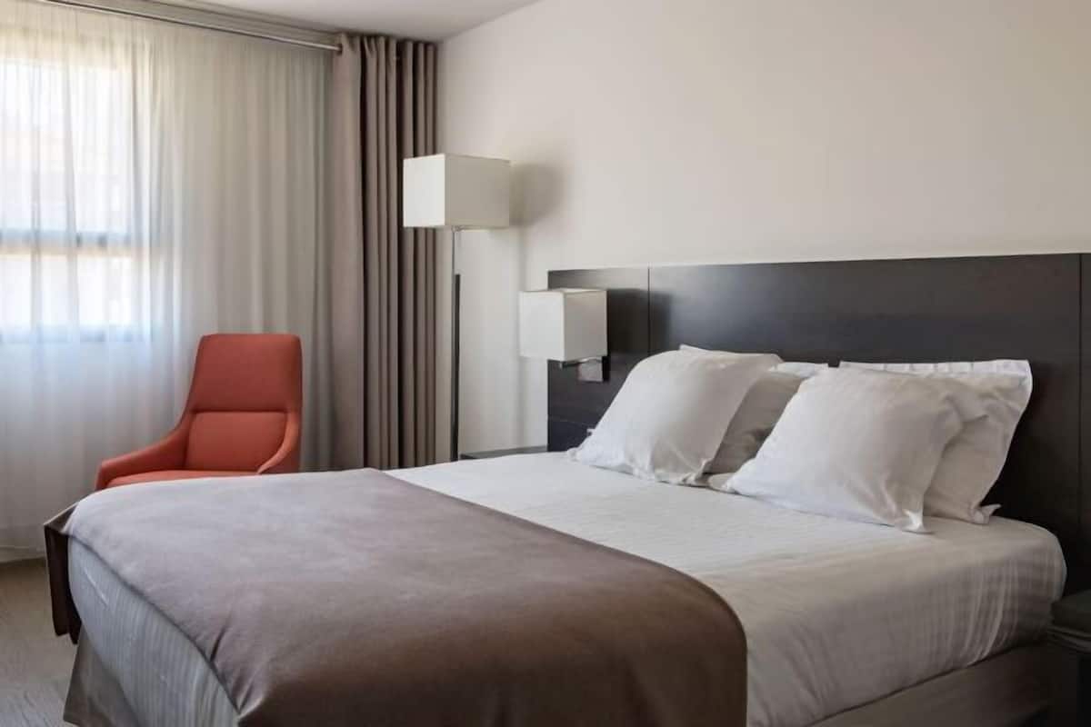 Hotellihuone, jossa on sänky, tuoli, yöpöytä, lamppu ja ikkuna, jossa on verhot.