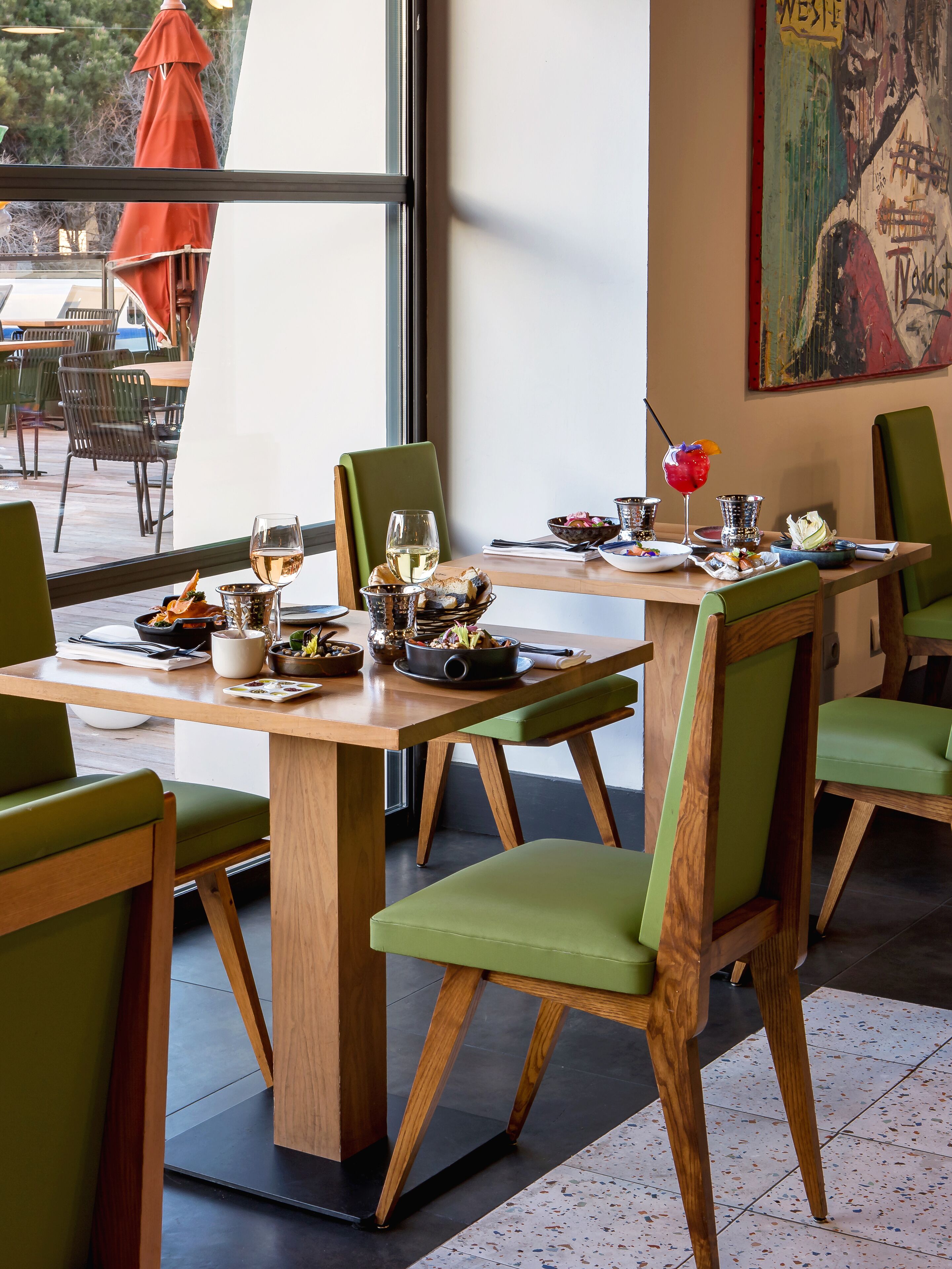 A restaurant interior with wooden tables and green chairs, a large window, and a painting on the wall.