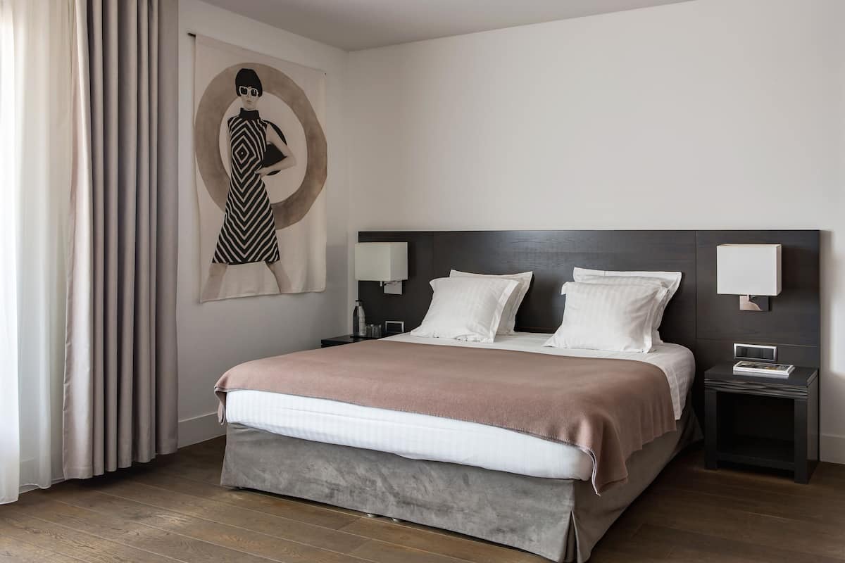 Moderni hotellihuone, jossa on suuri sänky, kaksi yöpöytää, yölamppu, seinälle ripustettu taideteos ja ikkuna, jossa on verhot.