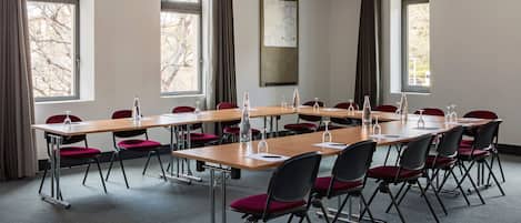 A conference room with long tables and red chairs, large windows with curtains, and a whiteboard on the wall.