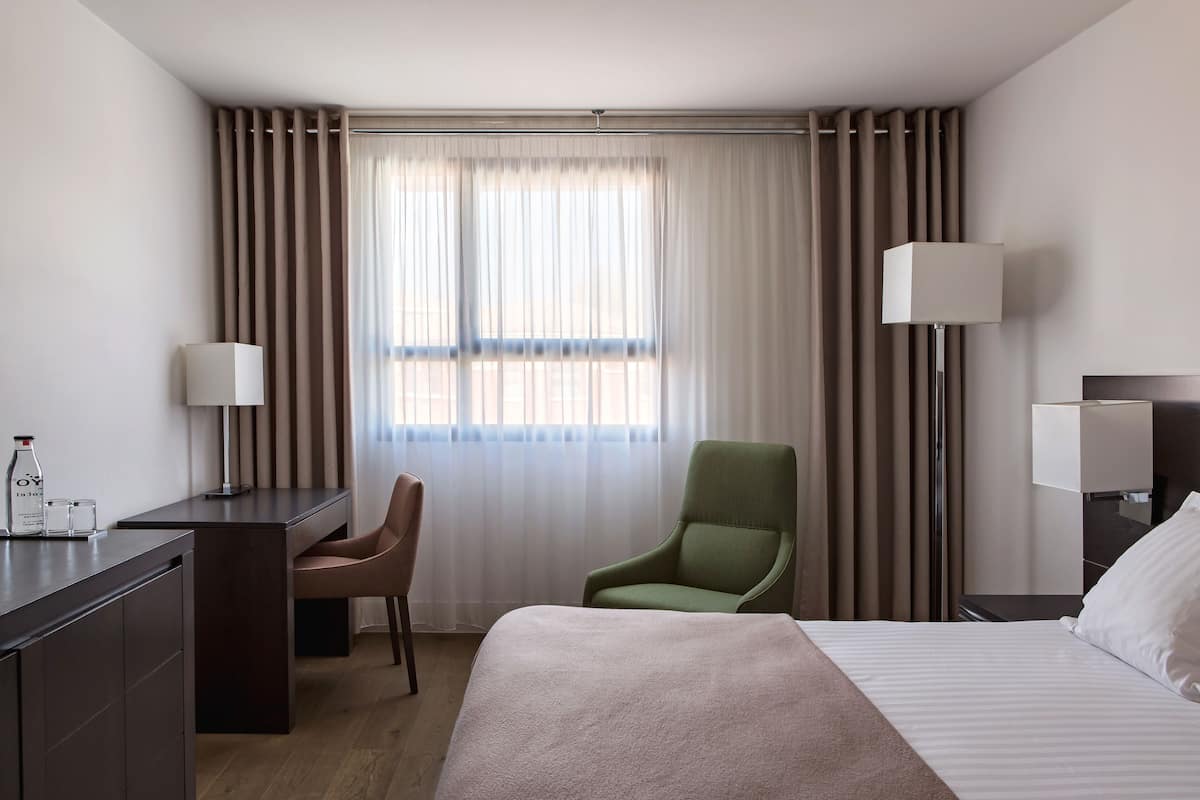 Hotellihuone, jossa on sänky, työpöytä, tuoli, lamppu ja ikkuna, jossa on verhot.