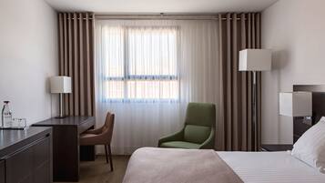 Une chambre d’hôtel avec un lit, un bureau, une chaise, une lampe et une fenêtre avec des rideaux.