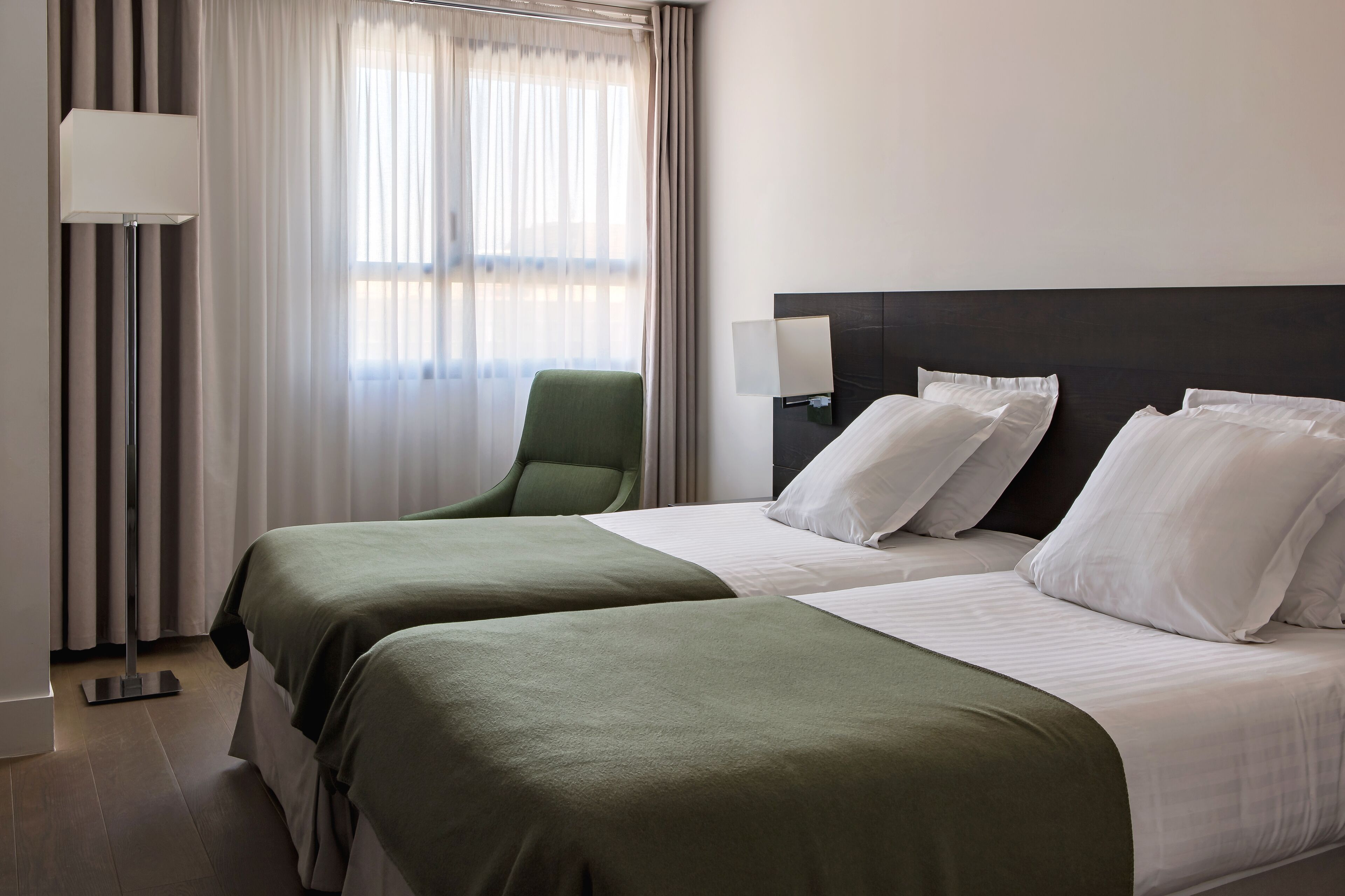 Habitación de hotel con dos camas, una silla verde, una lámpara y una ventana con cortinas.