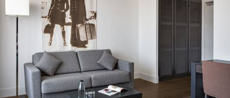 Una sala de estar moderna con un sofá gris, una mesa de centro negra con una botella y vasos, una lámpara de pie y un cartel grande en la pared.