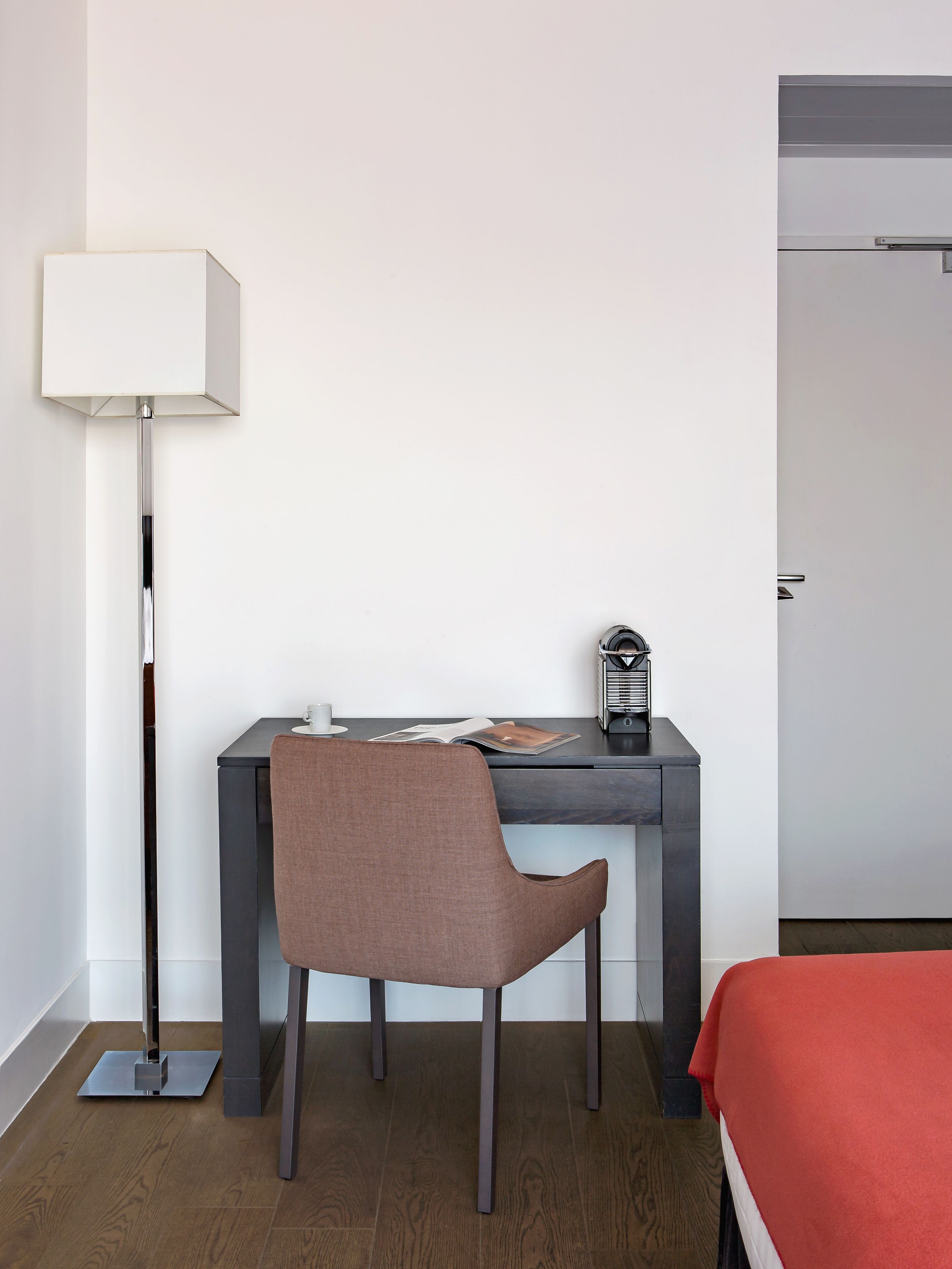 A hotel room with a desk, chair, lamp, and a bed with a red blanket.