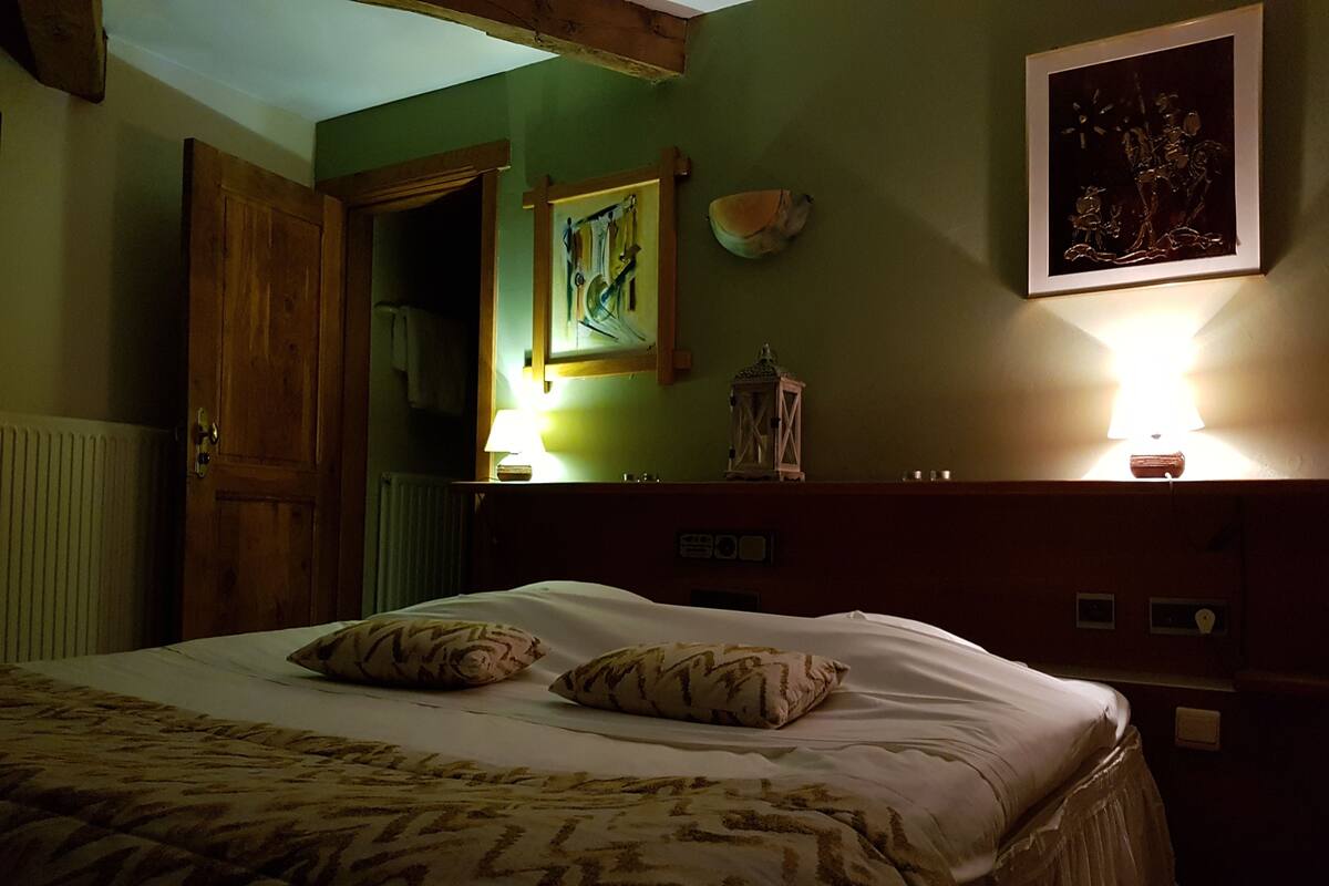 A bedroom with a bed, pillows, a nightstand, a lamp, a painting, and a wooden door.