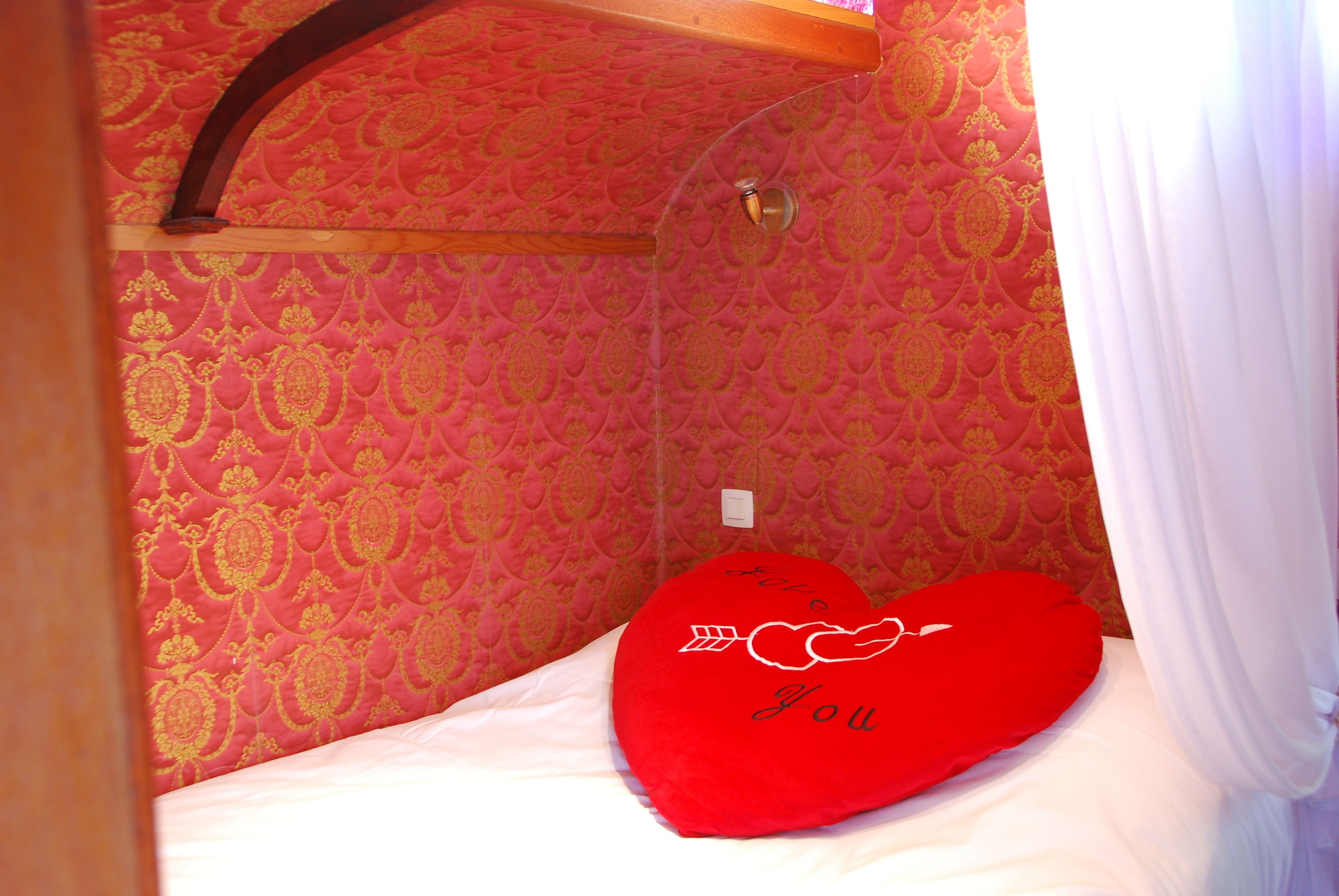 Een kamer met een bed, een rood hartvormig kussen met de tekst 'I love you' en behang met een patroon.