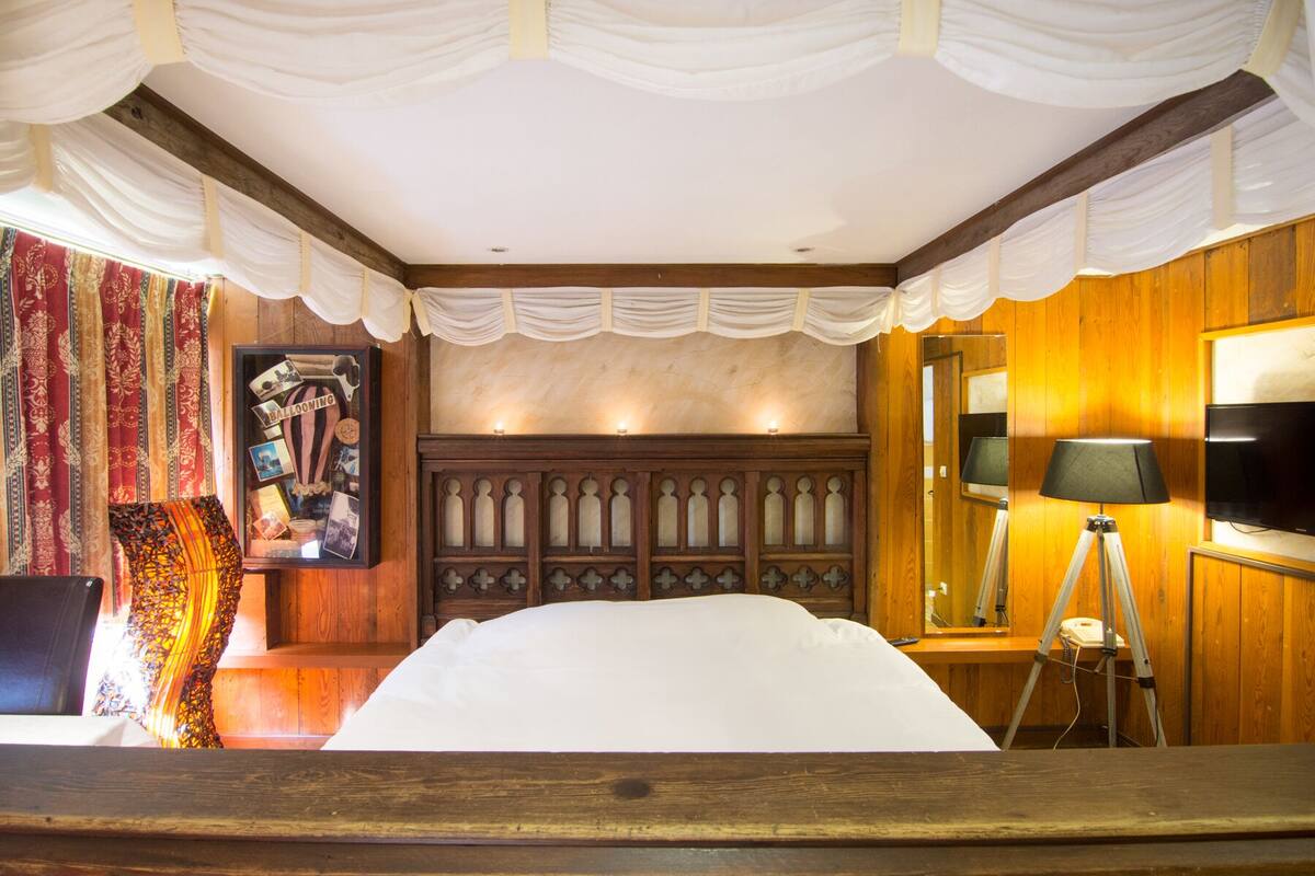 A bedroom with a wooden headboard, a bed with white bedding, a lamp, a television, and a wall with a framed photo and a decorative item.