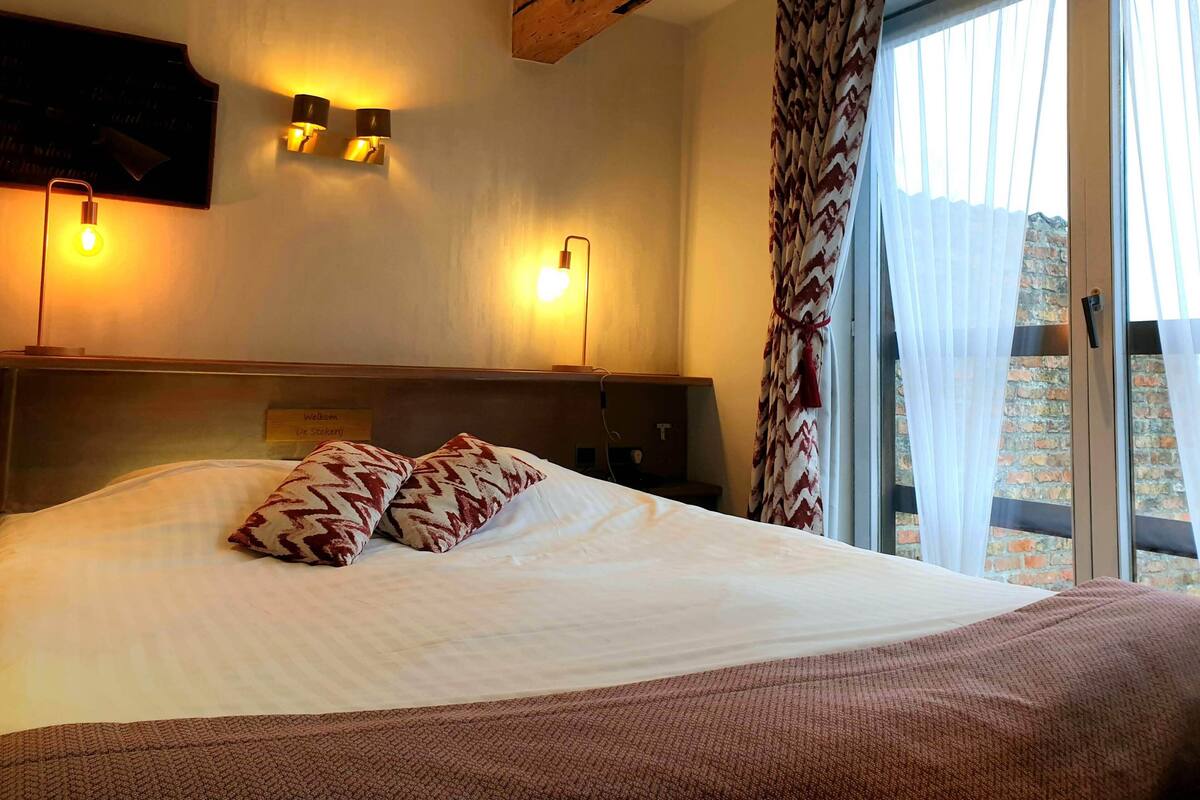 A hotel room with a large bed, two wall-mounted lights, a wooden ceiling beam, and a view of a brick building through a window with sheer curtains.