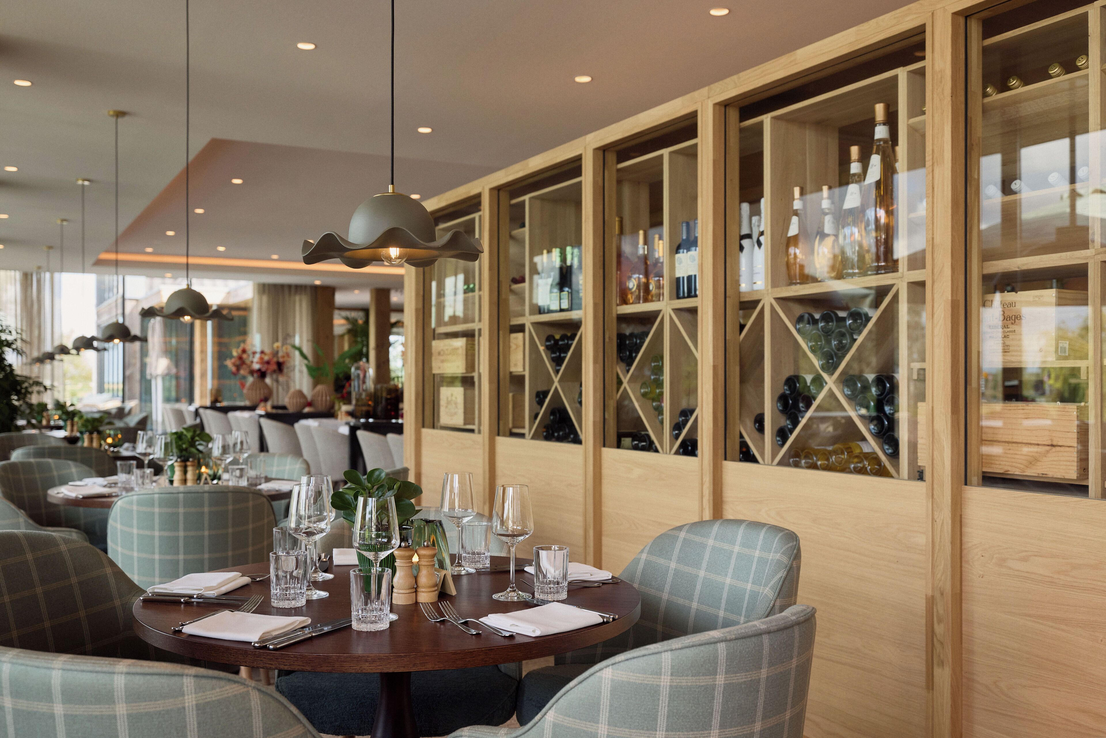 Un restaurant avec des tables rondes dressées pour le dîner, des étagères à vin en bois en arrière-plan et un éclairage moderne suspendu au plafond.