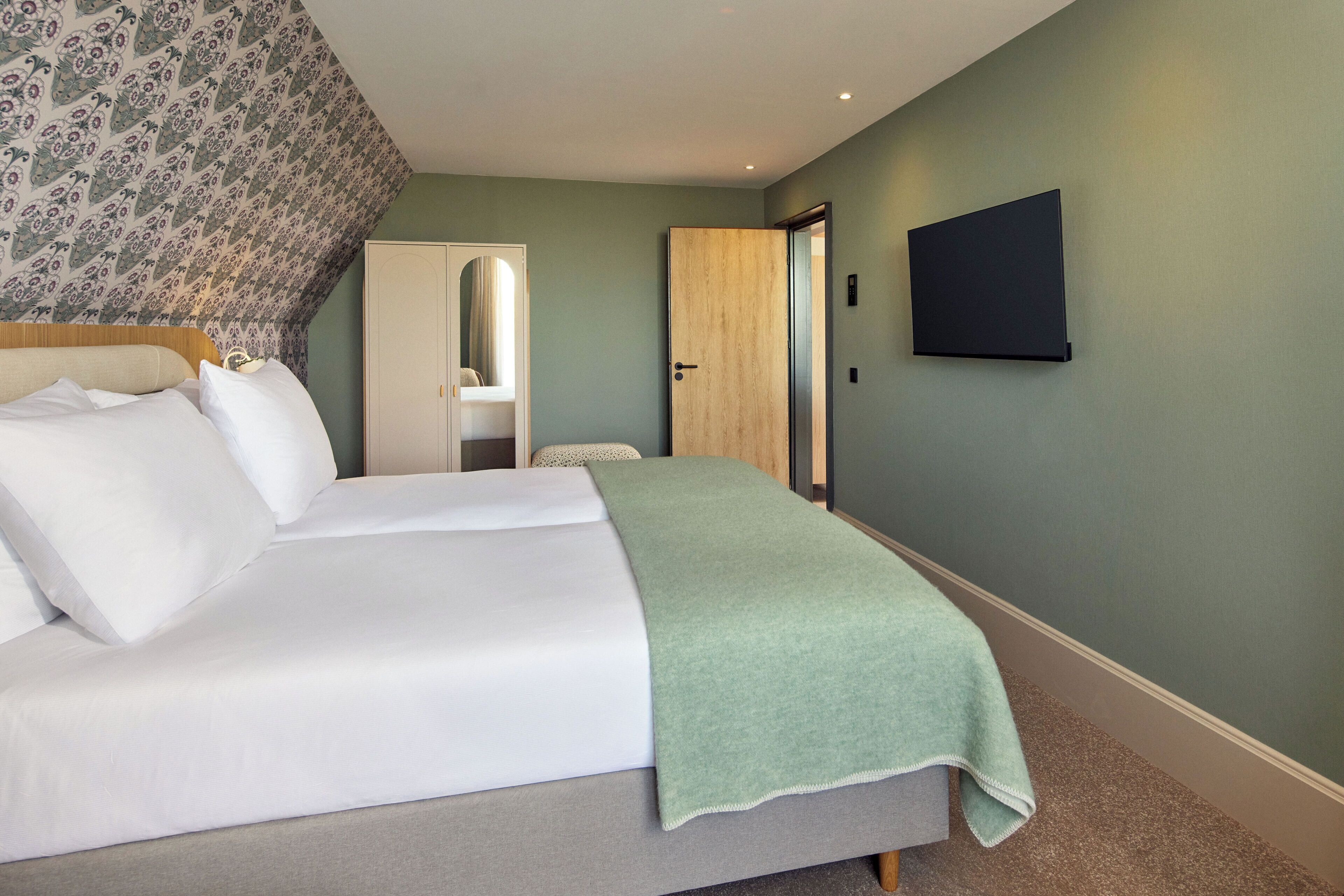 A bedroom with a bed, a green blanket, a wooden door, and a wall-mounted TV.