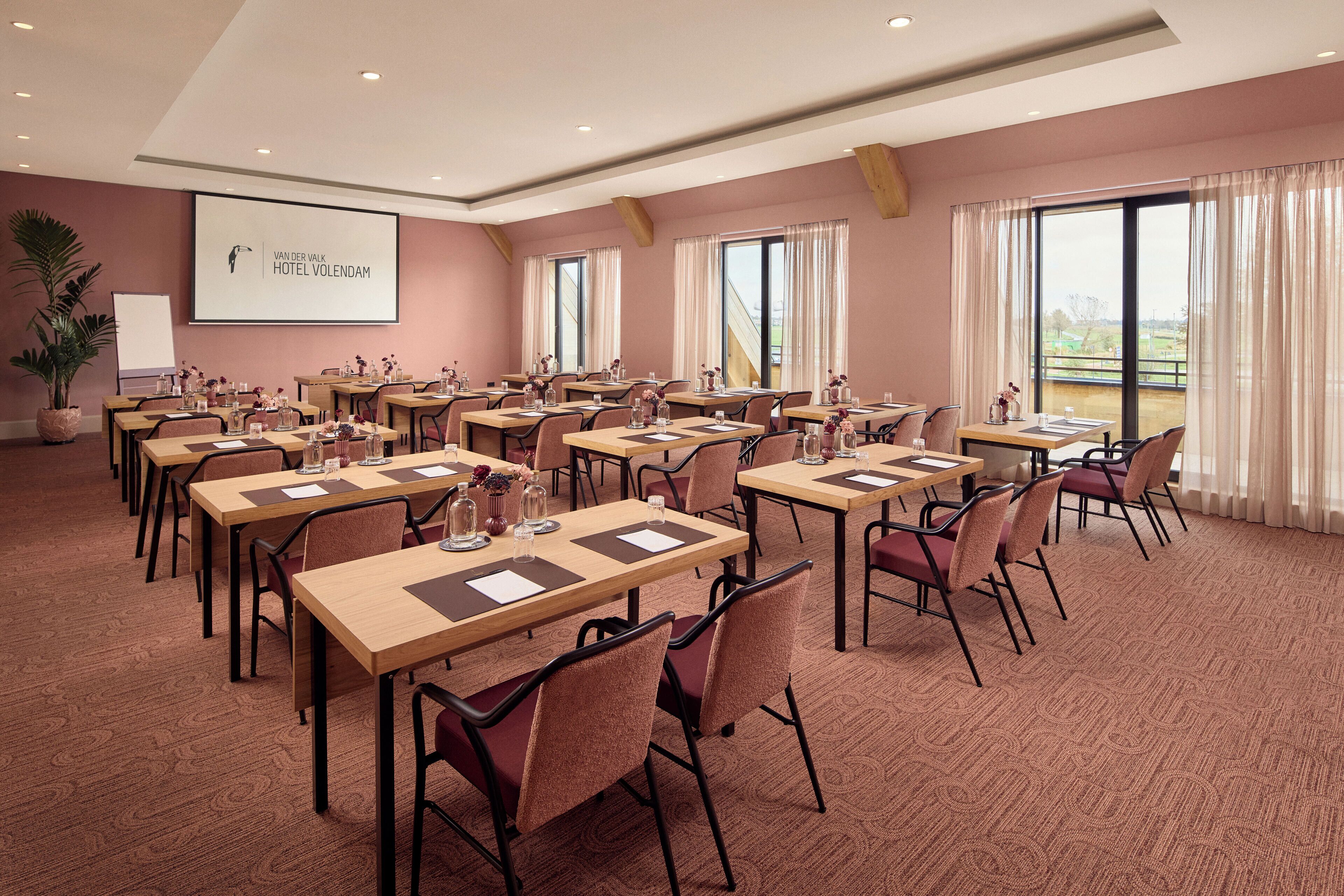 A conference room with multiple tables and chairs, a projector screen, and large windows with curtains.