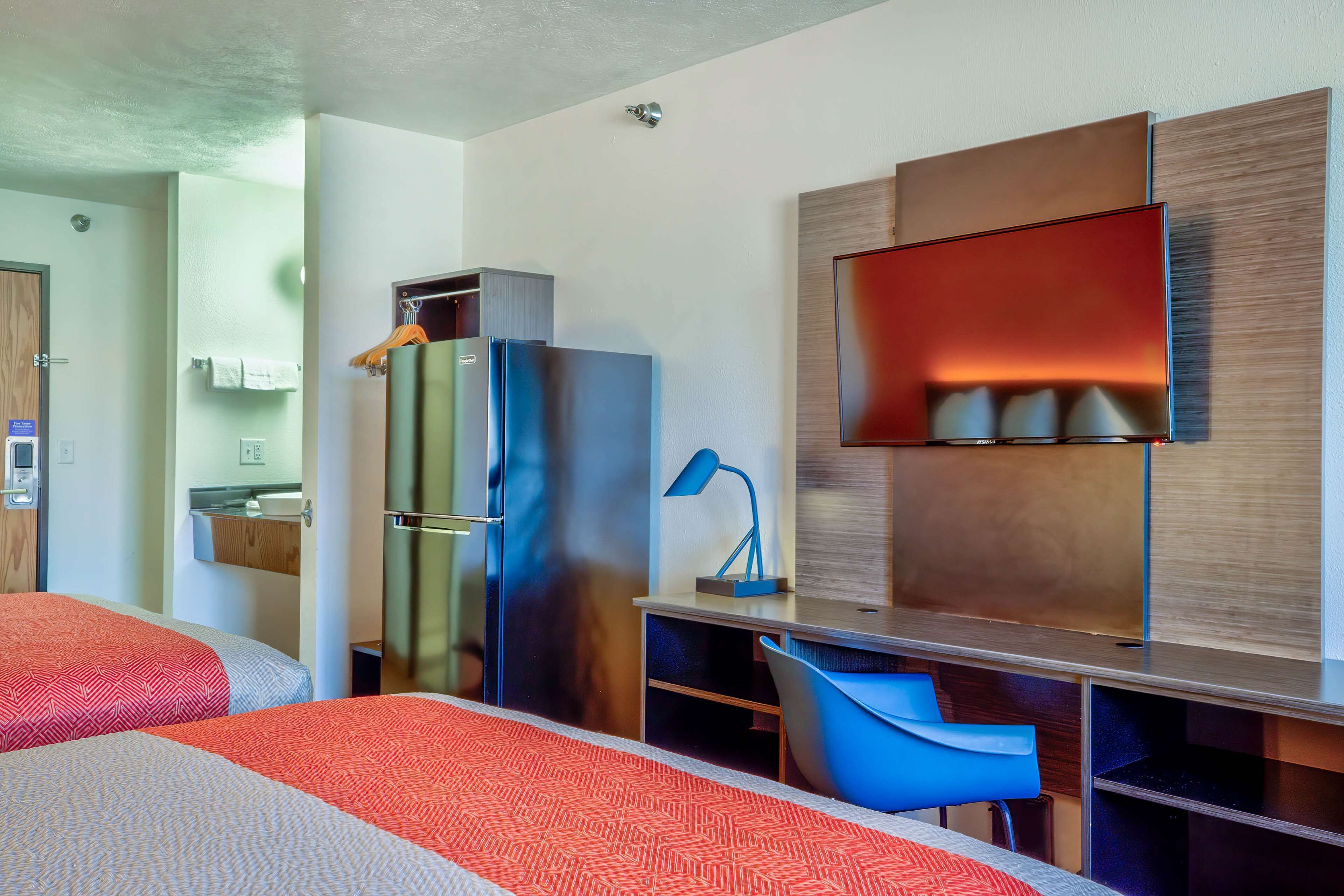 Ein modernes Hotelzimmer mit zwei Betten, einem Kühlschrank, einem Schreibtisch mit Stuhl, einem Fernseher und einem im Hintergrund sichtbaren Badezimmer.