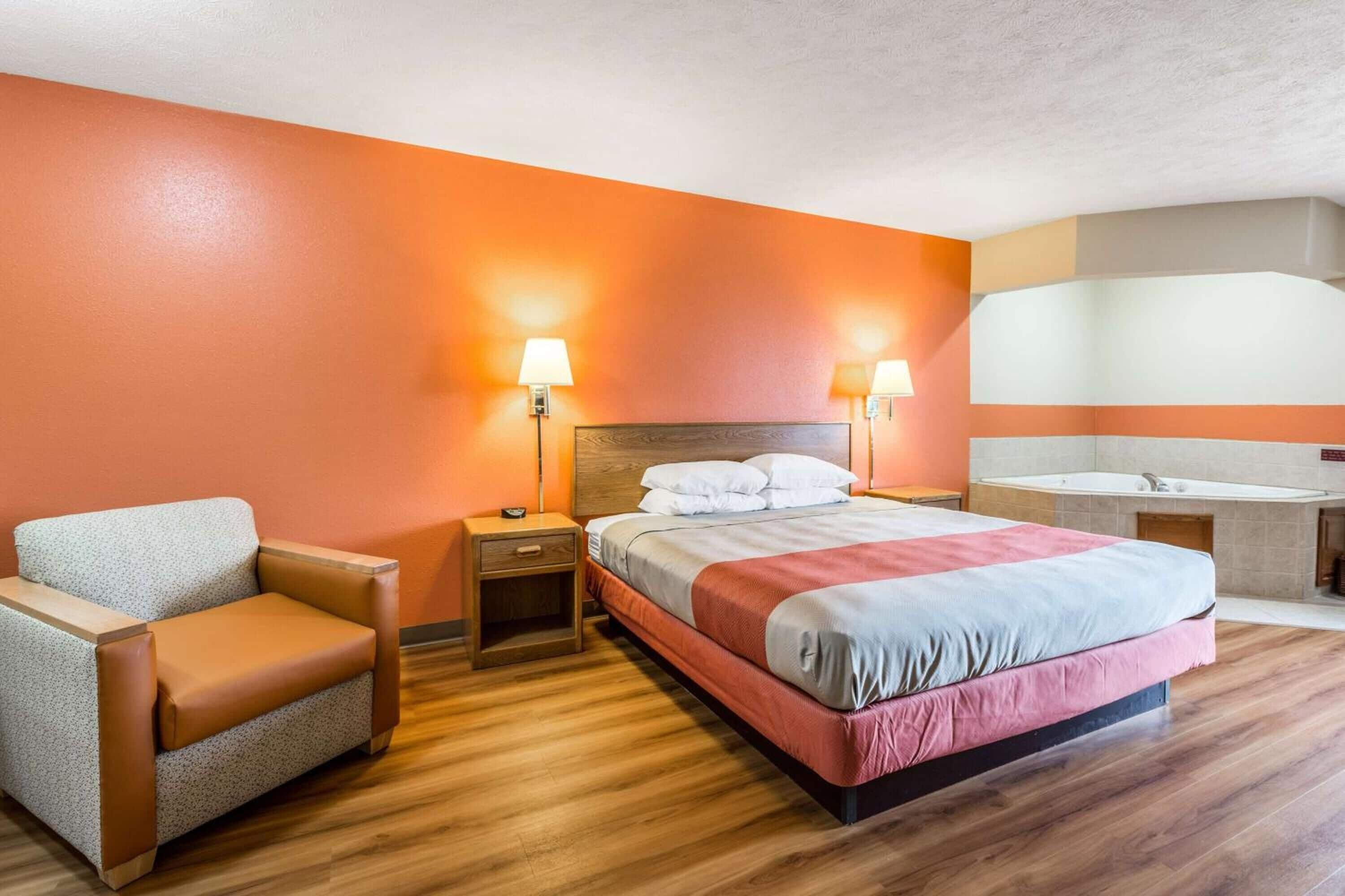 A hotel room with a bed, a chair, a nightstand, a lamp, and a bathroom with a sink and mirror.