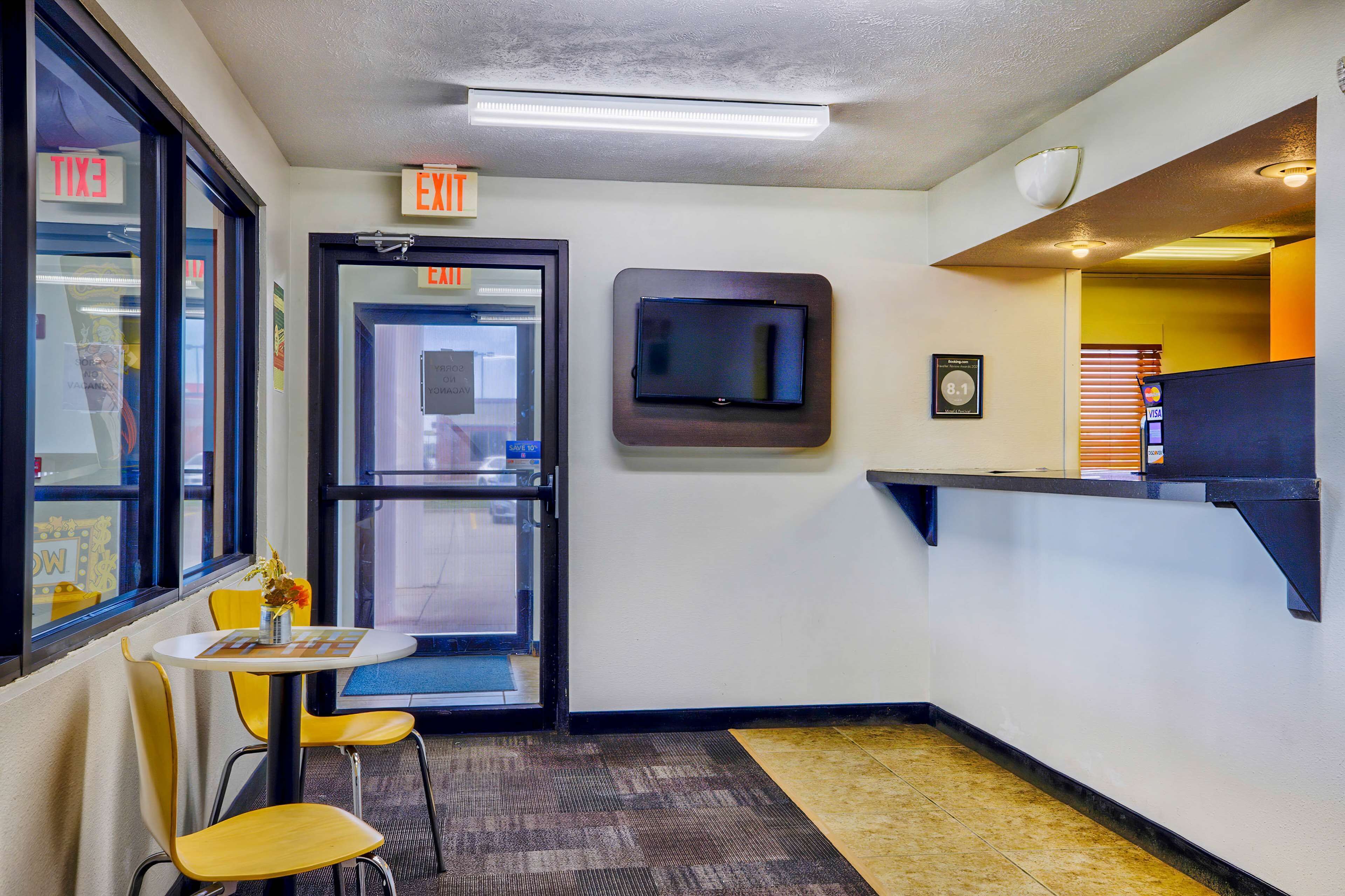 A small, brightly lit room with a single table and two chairs, an exit sign, a television mounted on the wall, and a vending machine.