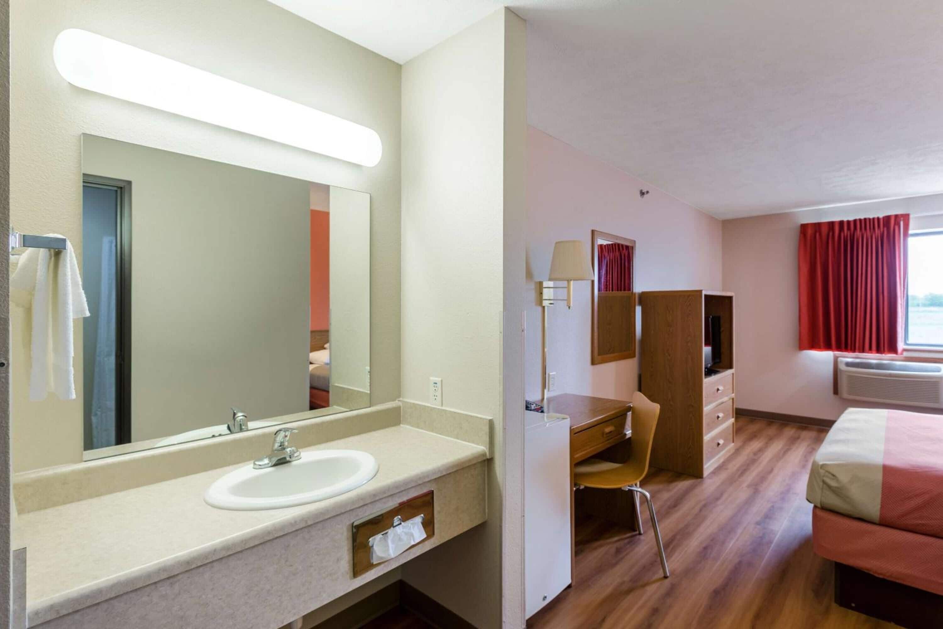 A hotel room with a large mirror, a sink, a desk, a chair, a bed, and a window with curtains.